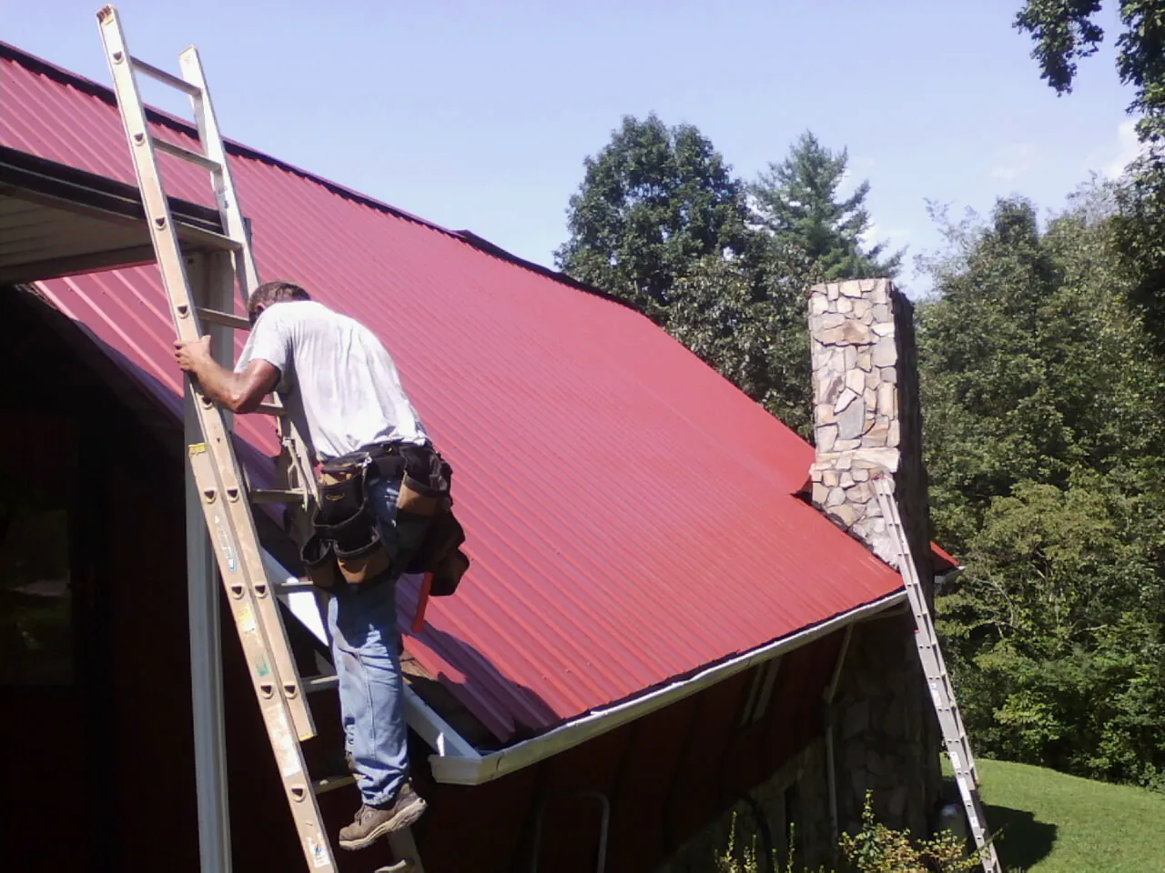 Roofer on a ladder beside a red metal roof and stone chimney — Soffit & Fascia Repair by Roofing Brothers in Yankton